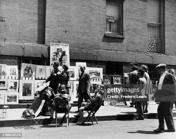 Two people sit on folding chairs as pedestrians inspect artworks on display at a sidewalk art exhibition in the Greenwich Village neighbourhood of...