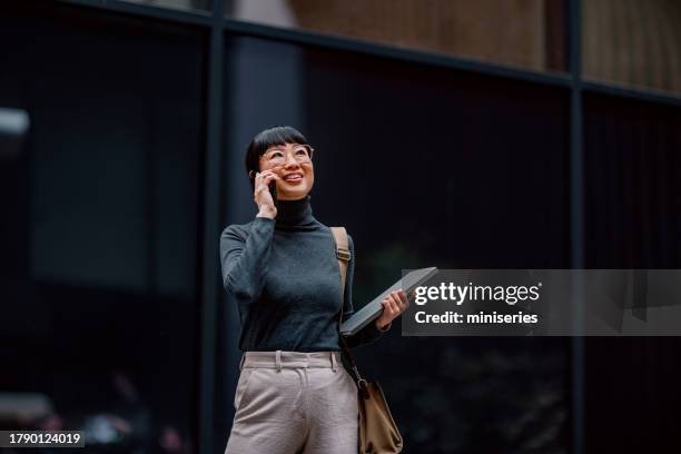 donna d'affari giapponese sorridente moderna che fa una telefonata mentre cammina in città - business woman foto e immagini stock