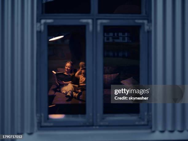 smiling mother reading a book to her daughter during night at home. - girl looking through window stock pictures, royalty-free photos & images