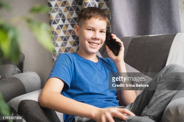 ritratto di un adolescente felice che parla su uno smartphone seduto a casa in salotto, guardando la fotocamera e sorridendo. - boy pre adolescent phone hand foto e immagini stock