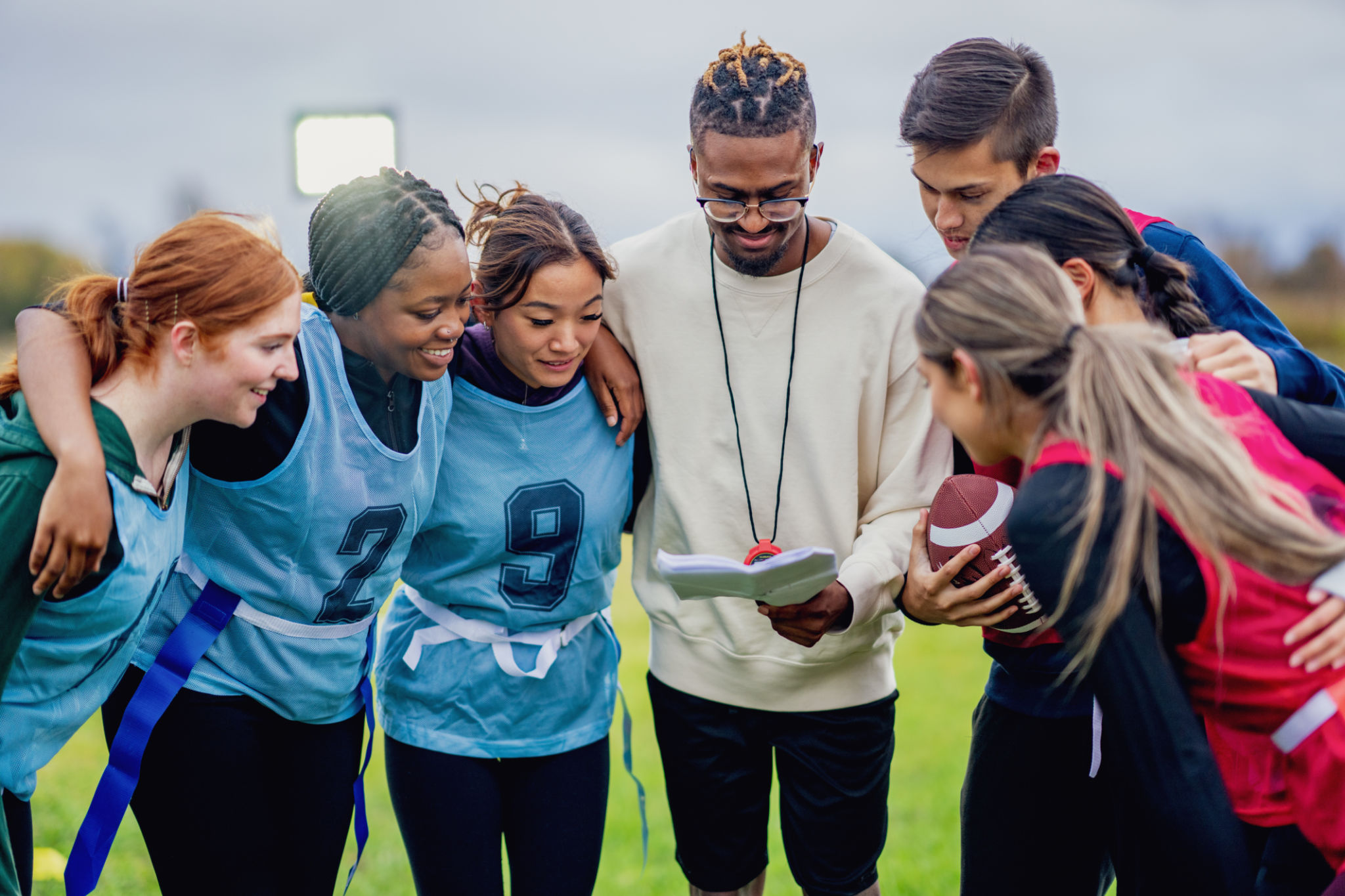 A small group of university students huddle in closely as they take instructions from their coach during a game of flag football. They are each dressed comfortably and wearing numbered team pinnies as they listen carefully to their coaches instructions.