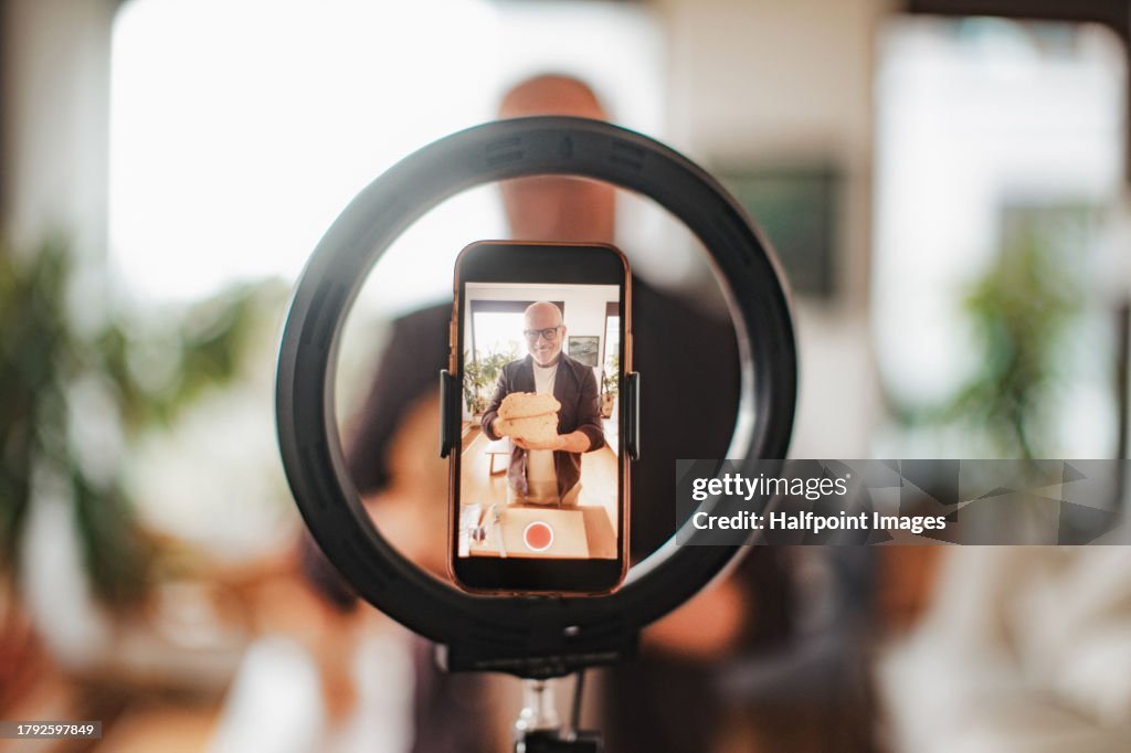 Senior food influencer making online bread baking course. Granfluencer preparing a bread, showing, explaining baking technique to the camera. Elderly man creating online content about baking, cooking and food for followers on social media.