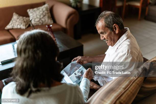 senior man talking with his wife while looking a photo album in the living room at home - vintage photograph stock pictures, royalty-free photos & images