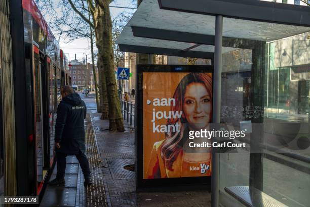Man boards a tram next to a People's Party for Freedom and Democracy campaign poster featuring a picture of party leader Dilan Yesilgoz-Zegerius on...