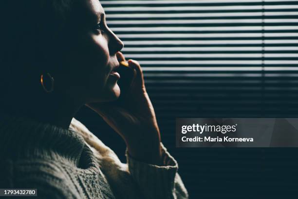 tense woman looks through the blinds while talking on the phone. - white woman calling 911 stock pictures, royalty-free photos & images