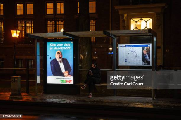 Woman checks her phone as she waits at a tram stop next to a campaign poster for the GroenLinksPvdA alliance featuring a picture of party leader...