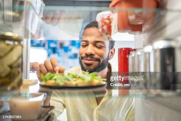 begeisterter junger schwarzer mann, der lebensmittel in seinem kühlschrank betrachtet - full fridge stock-fotos und bilder