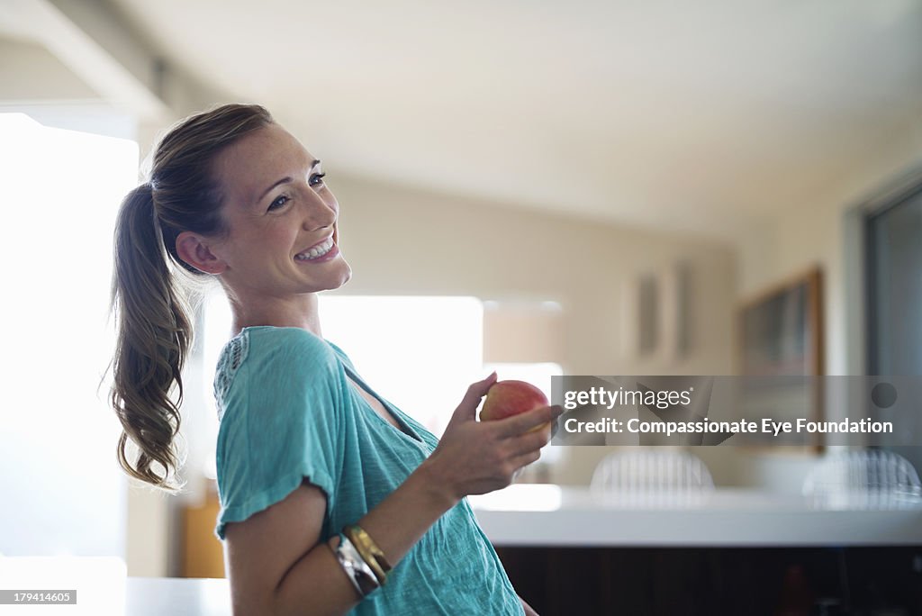 Smiling woman eating apple in kitchen