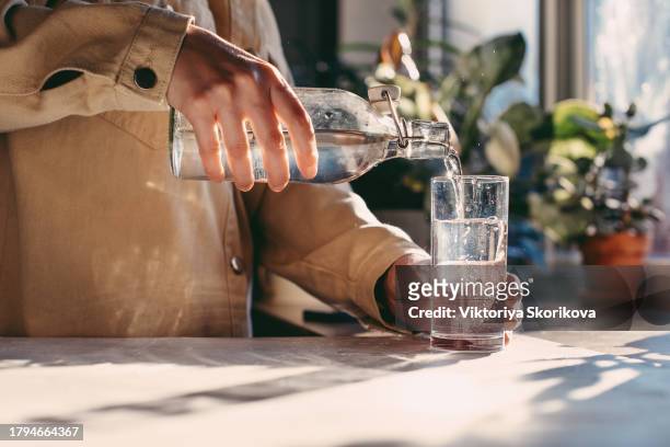 close up of woman pouring fresh water into glass from jug at home. attractive girl drinking water on domestic kitchen. young woman holding glass of water with lemon on kitchen in slow motion - girl filling water glass stock pictures, royalty-free photos & images