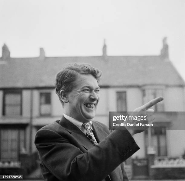 Author Kingsley Amis smiles while pointing in the street outside his house at 24 The Grove, Uplands, Swansea, Wales, 4th May 1956.