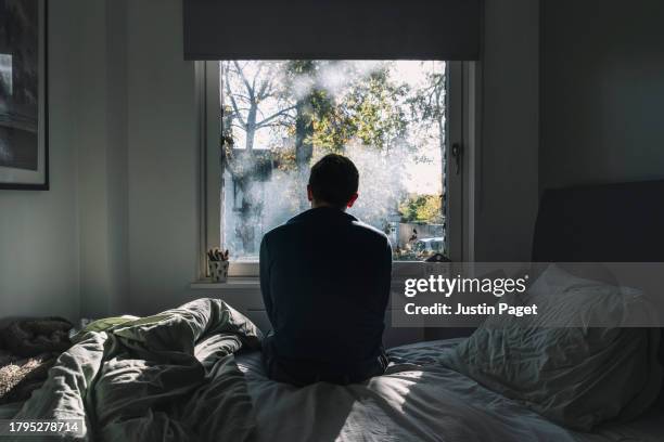 rear view of an unrecognisable man sitting on his bed looking out of the window - negative emotion - un solo hombre fotografías e imágenes de stock