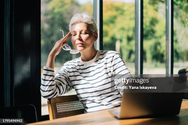 a middle-aged woman in glasses with short grey hair working with a laptop at her cafe, now closed her eyes, rubbing her temples, feeling tired. business owners - moe stockfoto's en -beelden