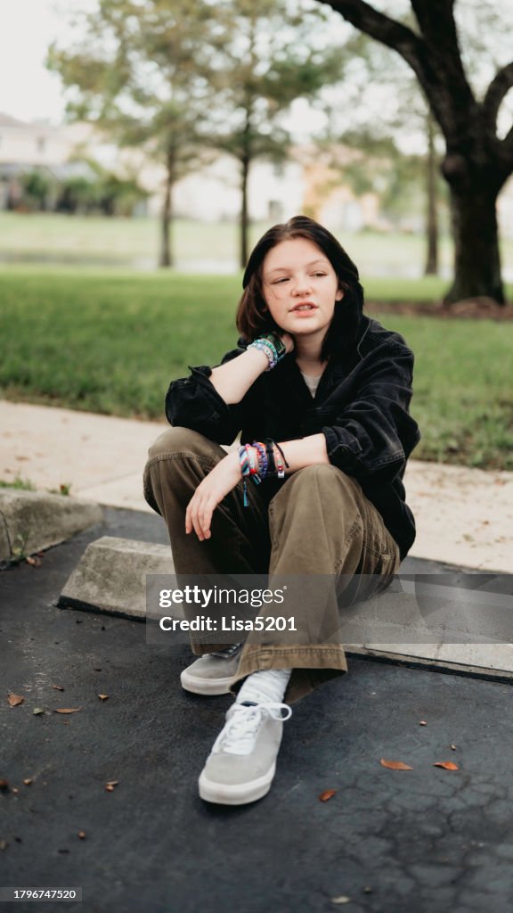 Transgender non-binary teen sits on a curb outdoors in casual moody serious portrait