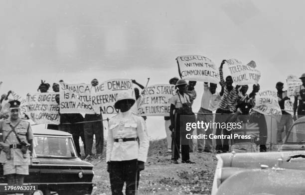 Police and military personnel stand before a group of protestors demonstrating with banners and placards as British Prime Minister Harold Macmillan...