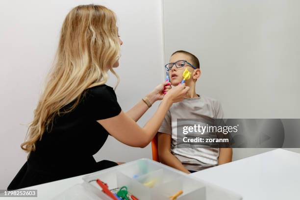 elementary age boy in occupational therapy, female doctor massaging his cheeks with special tools - speech pathology stock pictures, royalty-free photos & images