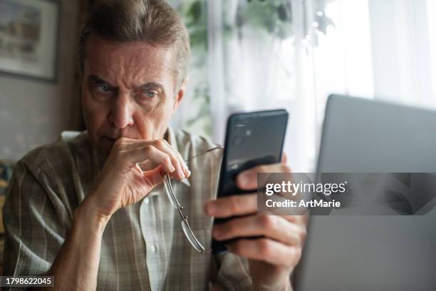 hombre mayor preocupado mirando el teléfono inteligente en casa - estafador fotografías e imágenes de stock