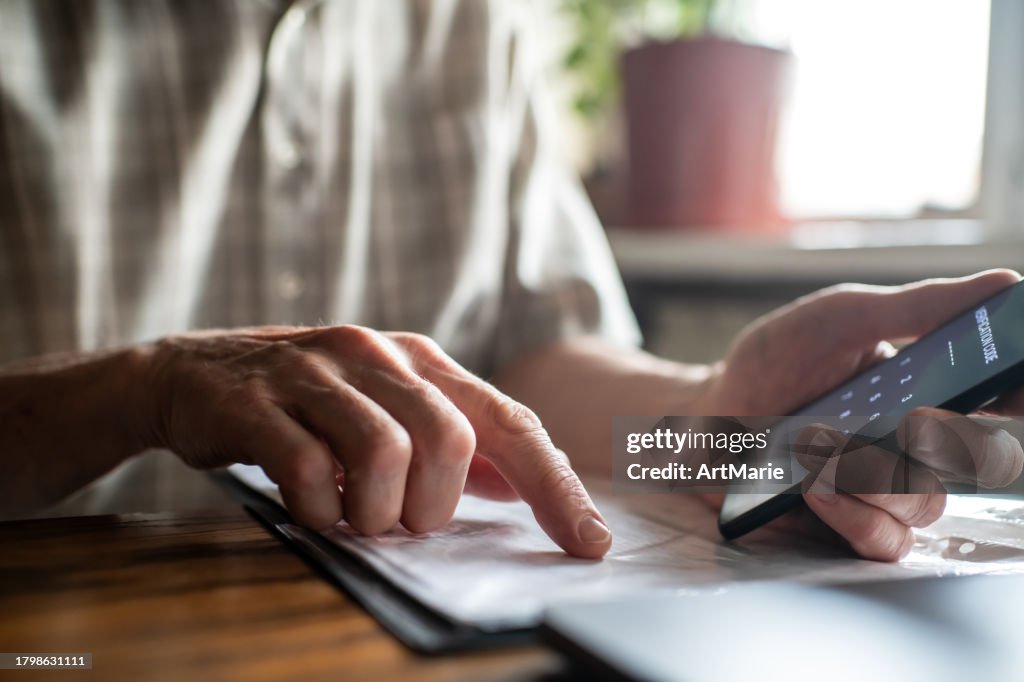 Senior man with smartphone, laptop and bills, documents or receipts at home enters verification code