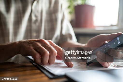 Senior man with smartphone, laptop and bills, documents or receipts at home enters verification code Senior man with smartphone, laptop and bills, documents or receipts at home enters verification code