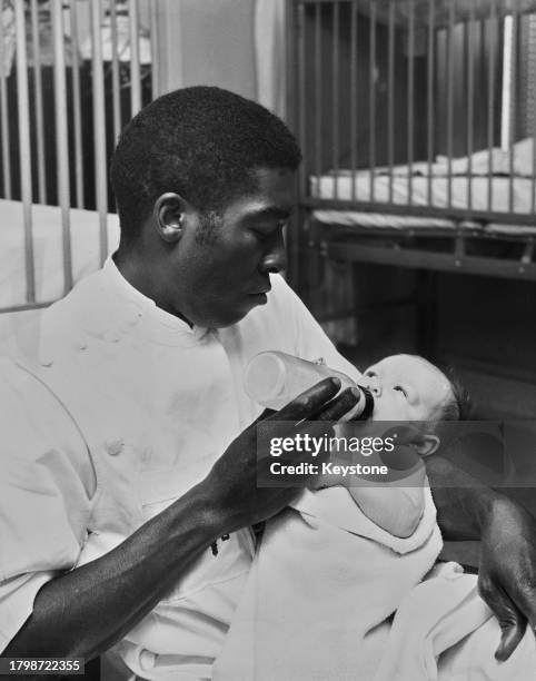 Nurse feeding a baby, his right arm supporting the child's head as his left hand holds the baby's bottle, at a hospital in Suffolk, United Kingdom,...