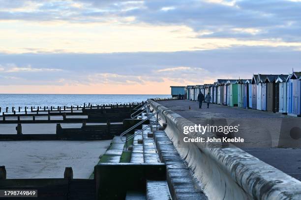 Couple walk their dog past beach huts on the beach at dusk on November 15, 2023 in Frinton-on-Sea, United Kingdom.