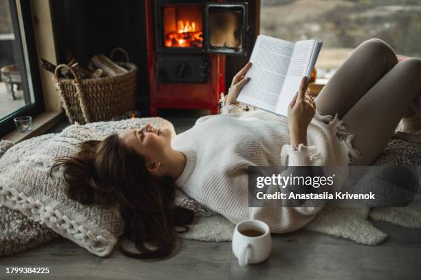 beautiful woman in warm clothes lying near the fireplace and reading book with beautiful view of wildlife from the window. - hygge stockfoto's en -beelden