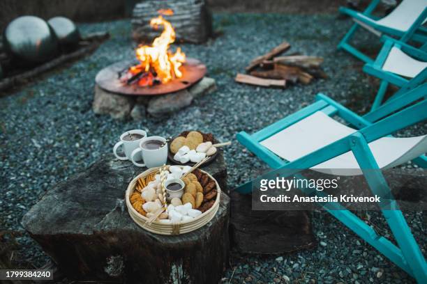 large outdoor fire pit surrounded by wooden chairs with mugs of hot cocoa, marshmallow, chocolate cookie and sweets - burnt cookies stock-fotos und bilder