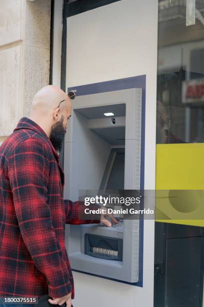 bohemian guy using an atm on a quiet downtown street, young man withdrawing cash from an atm. - loose change private view stock pictures, royalty-free photos & images