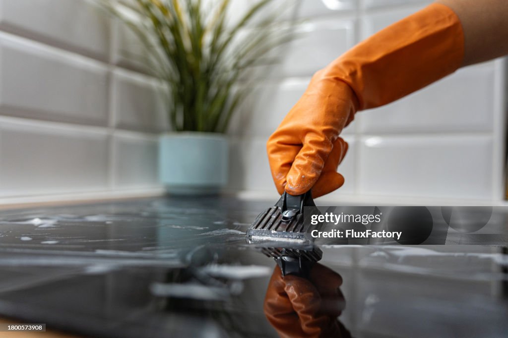 Close up of an unrecognizable woman cleaning the glass-ceramic stove top, while scarping the dirt from it