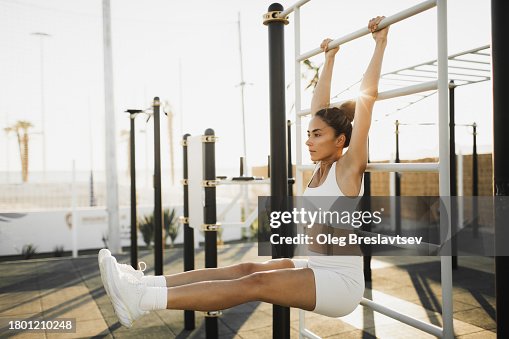 Athletic woman doing workout on horizontal bars, lifting legs to train abs muscles. Street fitness at sunset Athletic woman doing workout on horizontal bars, lifting legs to train abs muscles. Street fitness at sunset