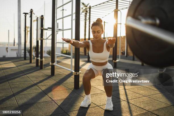 slim and beautiful sportive woman doing squats on outdoors sports ground at sunset - agachar se imagens e fotografias de stock