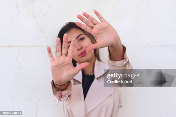 young woman wearing a trench coat, gestures with her palms and covers her face. - trench mouth stock pictures, royalty-free photos & images