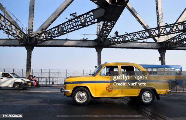 An iconic yellow ambassador taxi is driving past the Howrah Bridge in Kolkata, India, on November 26, 2023. These iconic taxis have been plying the...