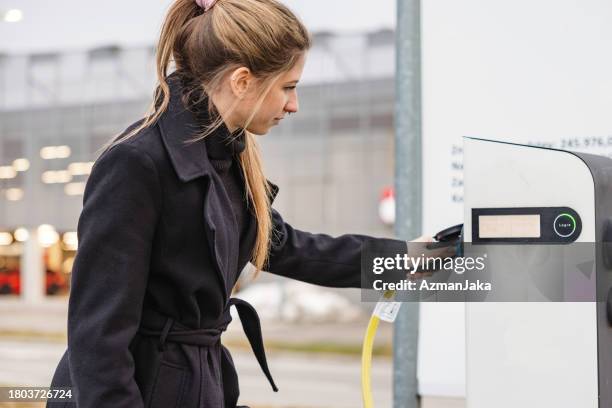 attraktive junge erwachsene frau, die sich darauf vorbereitet, ihr elektroauto an einer ladestation aufzuladen - hineinstecken stock-fotos und bilder