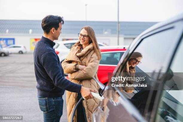 adult caucasian couple standing by a car and having a conversation while their electric car is charging - gas station friends stock pictures, royalty-free photos & images