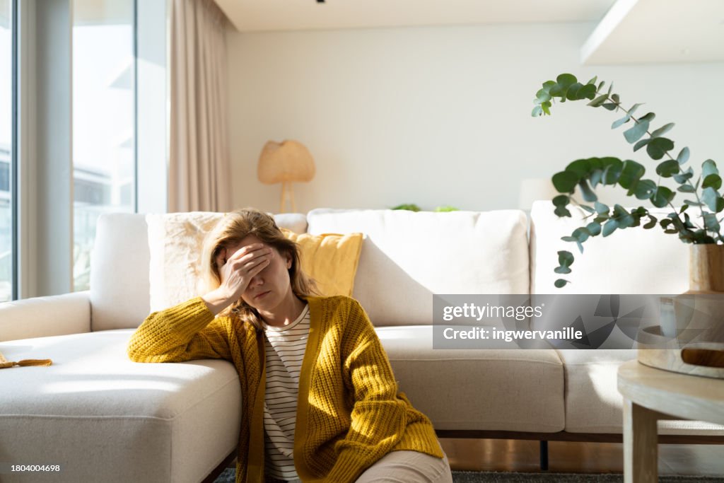 Woman having headache and holding her head while sitting on the floor at home
