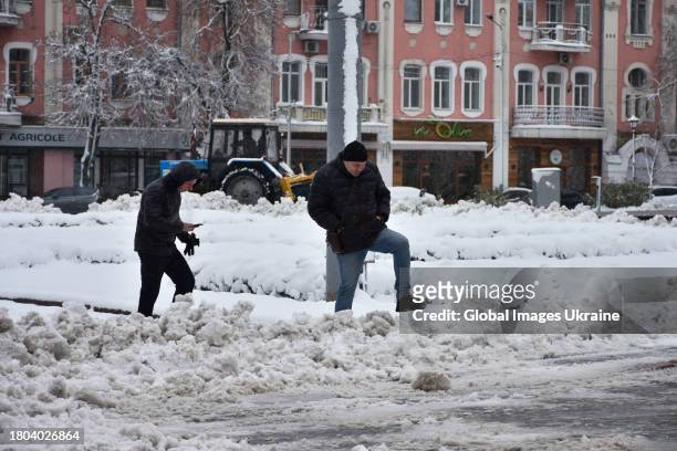 Men walk through the piles of snow, and behind them a tractor removes snow on a street on November 20, 2023 in Poltava, Ukraine. Since the snowfall...