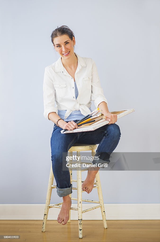 Studio Shot of woman sitting on stool, holding paintbrushes and artist's canvas