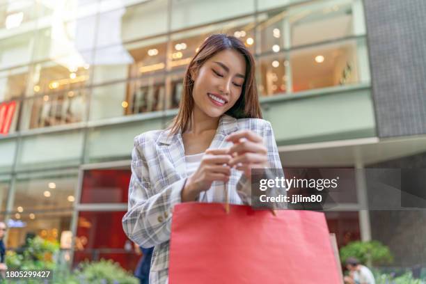 junge asiatische geschäftsfrau posiert nach einem erfolgreichen einkaufsbummel fröhlich mit einkaufstüten in einem einkaufszentrum und schwelgt in der einkaufstherapie. - shopping spree stock-fotos und bilder