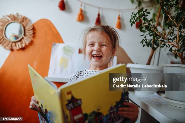emotional 5-year-old girl enjoys reading a book in her room at home. looking at camera - storytelling stock pictures, royalty-free photos & images