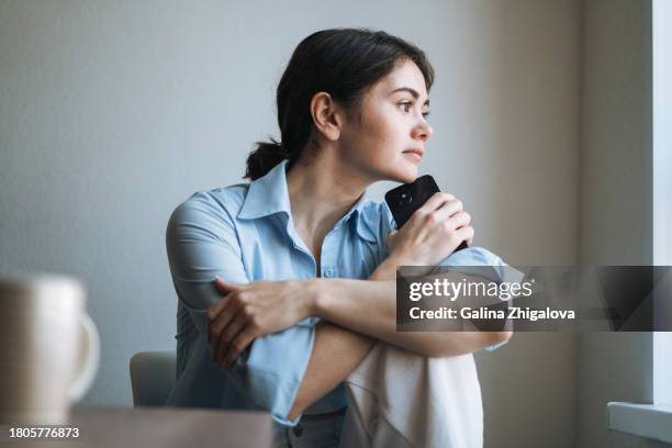 portrait of thinking young brunette woman with brunette hair using mobile phone near window at home - raccolto foto e immagini stock