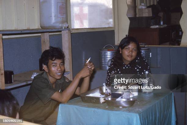 Uniformed man holding a cigarette, and a woman in civilian clothes, sit at a dirty table laden with metal pans, a syringe, and a large glass jar of...