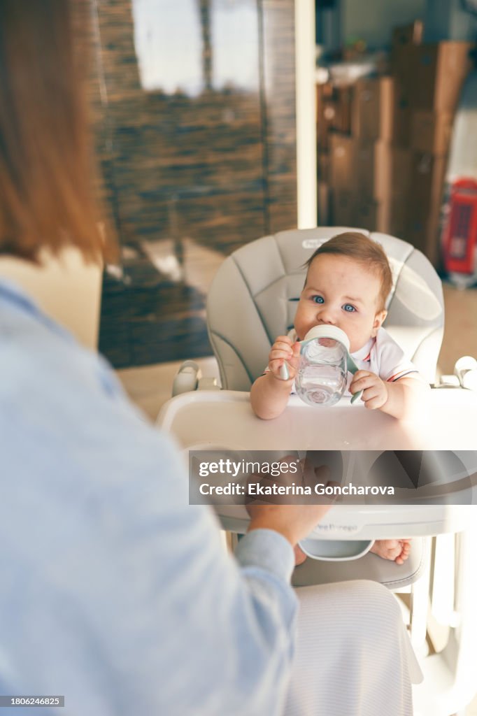Mother feeds the baby sitting at the feeding table in the kitchen in the morning