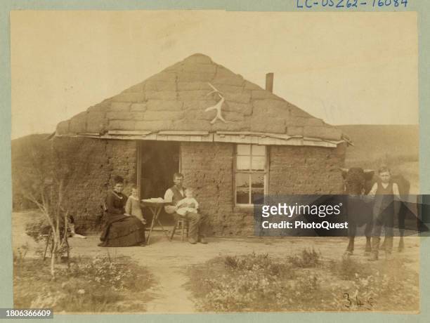 Portrait of an unidentified family in front of a sod house in Custer County, Nebraska, circa 1886. The parents, each with a child on their laps, sit...