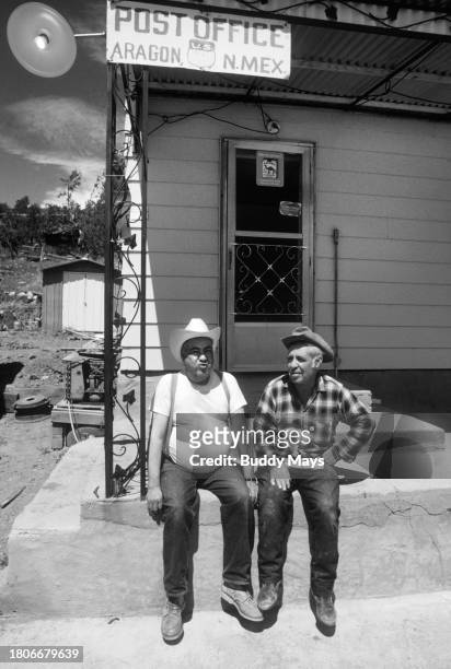 Two elderly Hispanic men, both og them retired farmers, sit on the steps of the U.S. Post Office in Aragon, New Mexico, chatting about the week's...