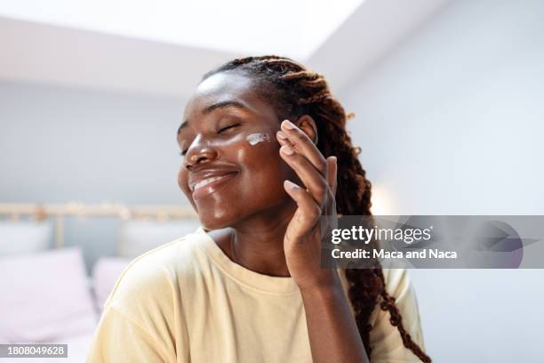 a close-up of a young african american woman cheerfully smiling, while applying beauty cream on her face - gezichtscrème stockfoto's en -beelden