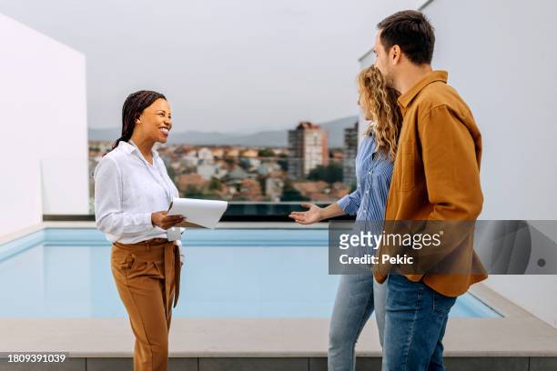 immobilienmaklerin im gespräch mit jungem, lässig gekleidetem paar am swimmingpool auf dem dach des gebäudes - rooftop pool stock-fotos und bilder