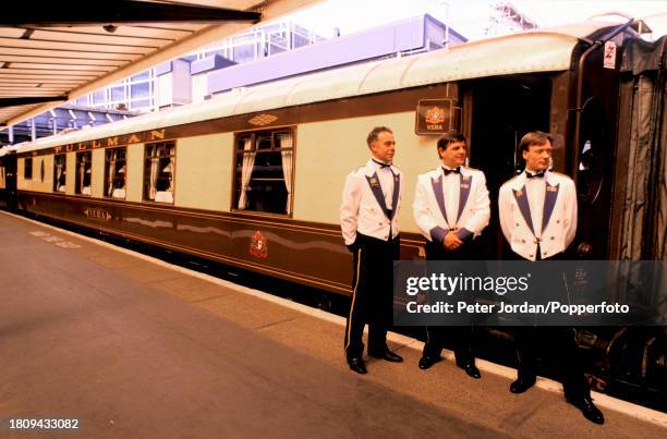 Waiters stand on a platform beside pullman dining cars of the Venice Simplon Orient Express luxury train service prior to departing from Victoria...