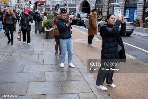 Man taking photographs on a digital rangefinder camera of a woman also taking pictures on her smartphone along Regent Street on 13th November 2023 in...