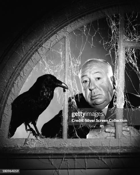 English director Alfred Hitchcock posing at a cobweb-strewn window with a stuffed crow, in a promotional portrait for the TV anthology series 'Alfred...