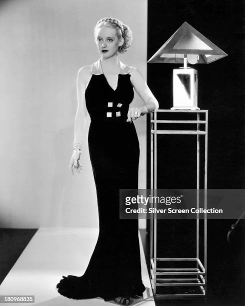American actress Bette Davis posing in a black, floor-length dress with contrasting collar and buttons, next to an Art Deco lamp and table, circa...